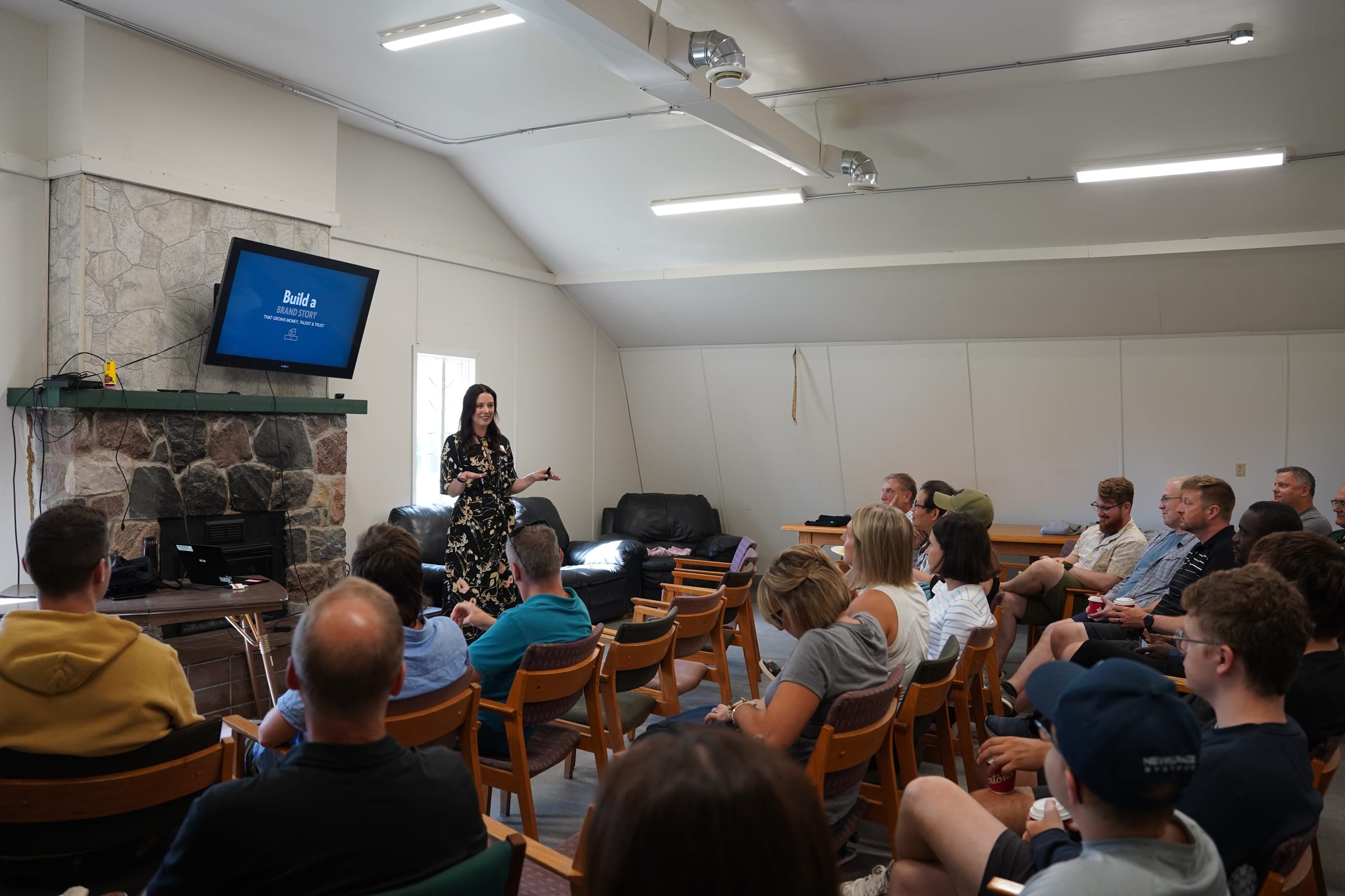 A presenter in a floral dress addresses a seated audience beside a stone fireplace, a screen reading 'Build a Brand Story' behind her, at a Co.Labs workshop.